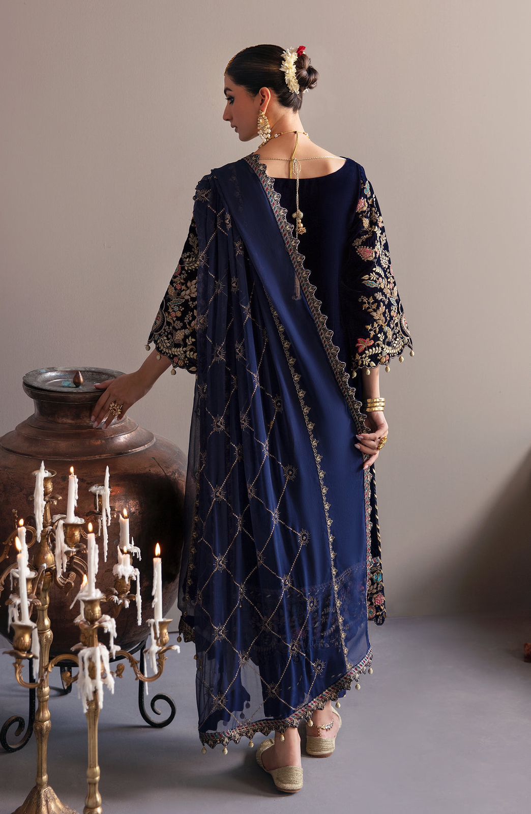 Emaan Adeel  Velvet Woman in a blue embroidered outfit standing next to a large vase and candelabra.