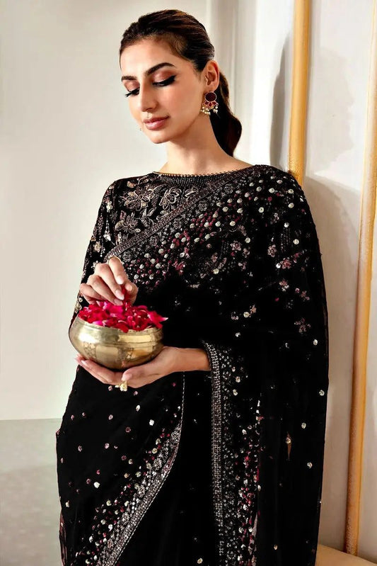 Woman in a black saree holding a brass pot with flowers indoors.