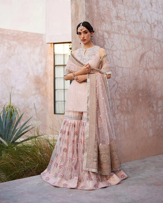 Faiza Saqlain Bridal Wedding Lehnga Garara Woman in a traditional outfit standing against a textured wall with plants.