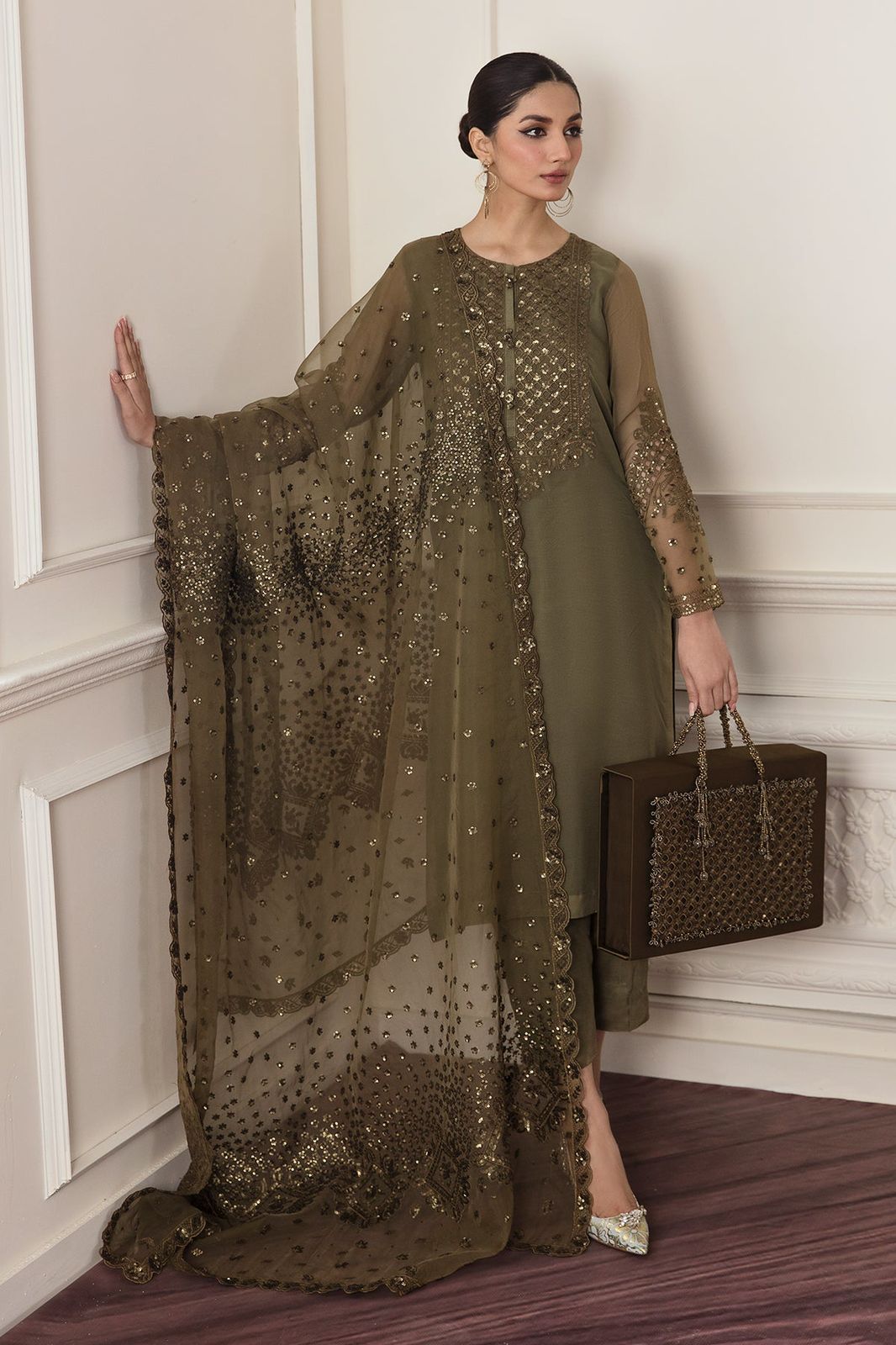 BAroque Formal Dress in a long, dark green dress with sheer overlay holding a matching handbag against a white wall.